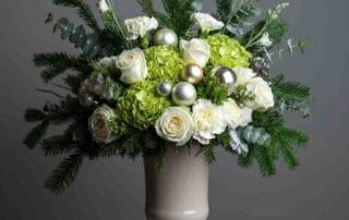 This centerpiece features a luxurious array of white roses, green hydrangea, white carnations mixed wintergreens and festive accents.